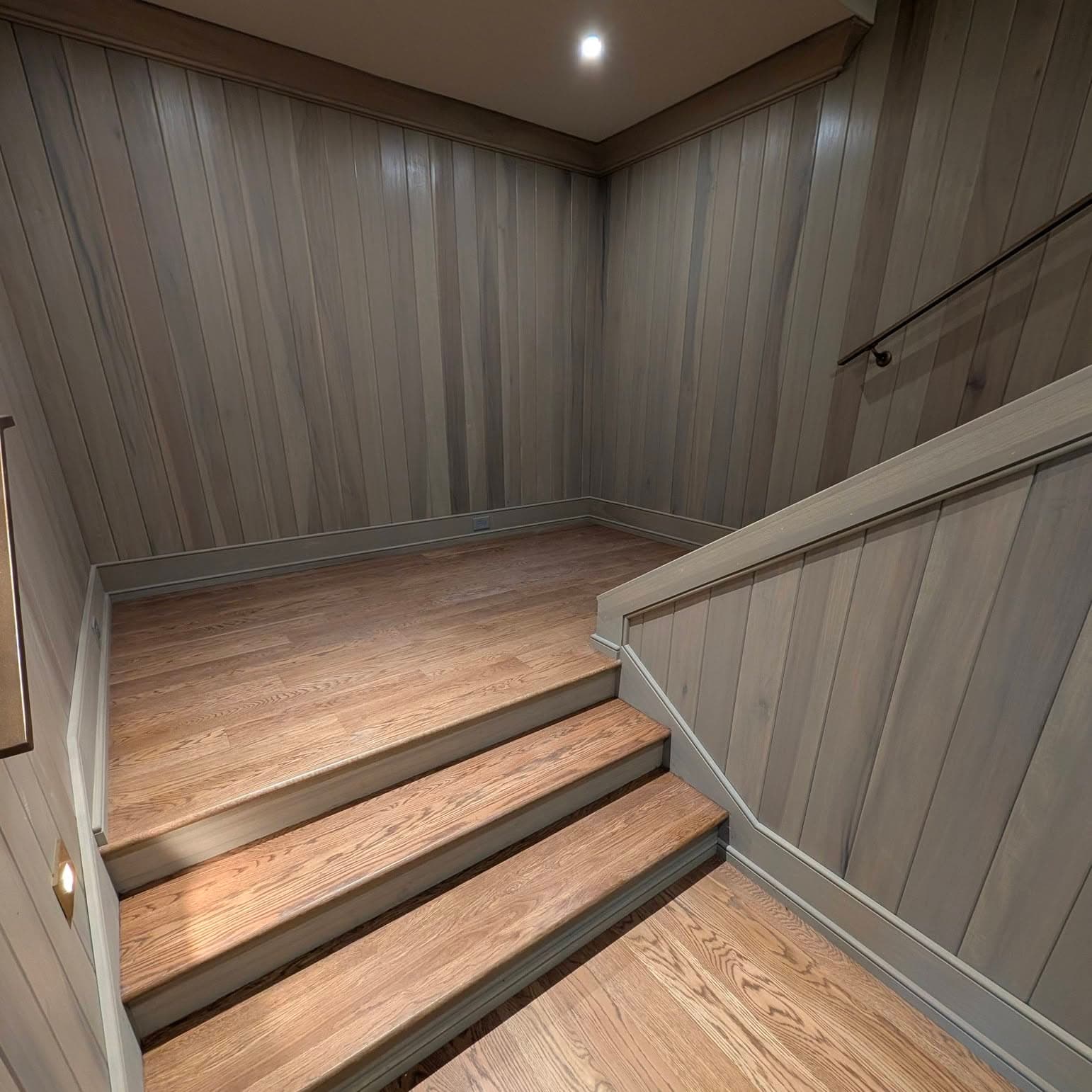 Stairs with dark risers into a wood-paneled stairwell, handrail on wall