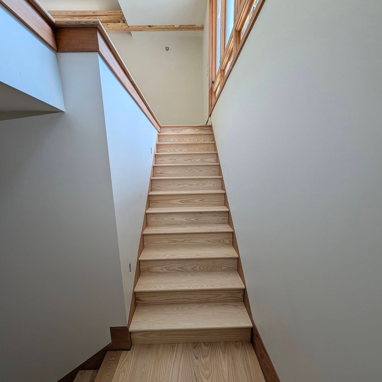 Straight light wood stair between two white walls, viewed from the base