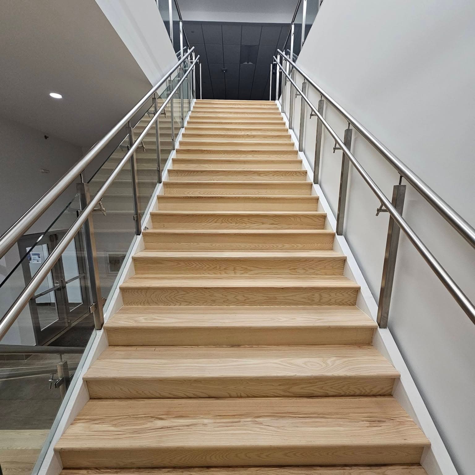 Looking up a long straight light wood stair with glass/metal railings both sides